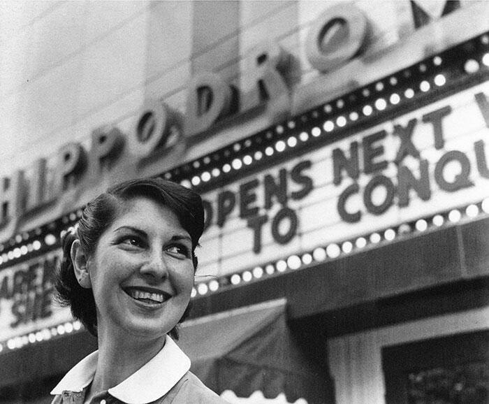 A woman stands smiling in front of a theatre marquis.