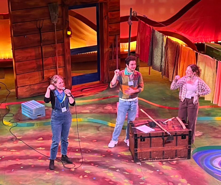 Three people standing on a stage with the backdrop of a house behind them, and a chest on the floor in front of them.