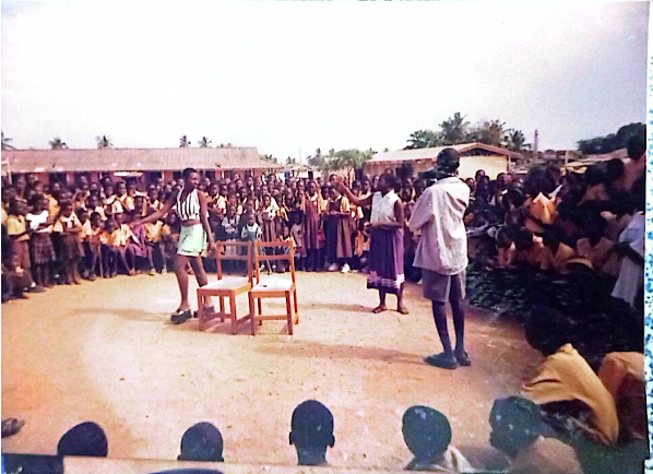 Three students stand with two chairs in the middle of a circle of audience members outside.