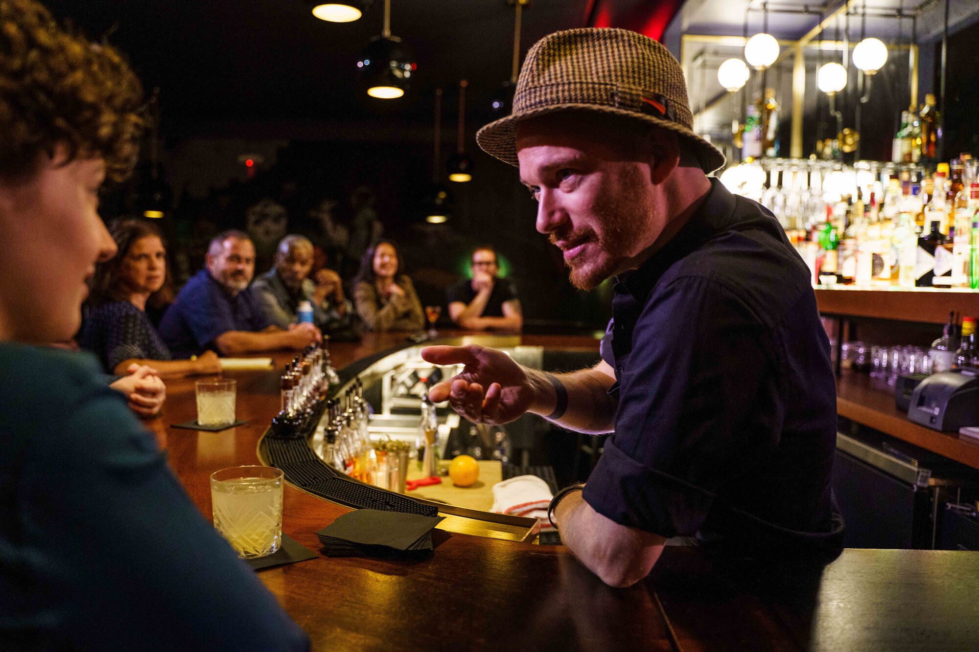 A bartender in a fedora speaks to a bar patron.