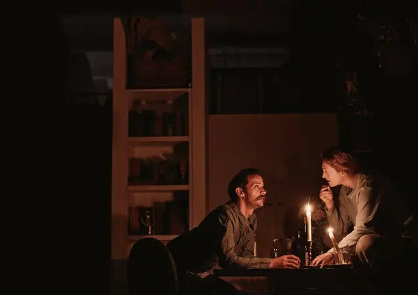 A man and a woman sit on either side of a candle on a dimly lit stage.