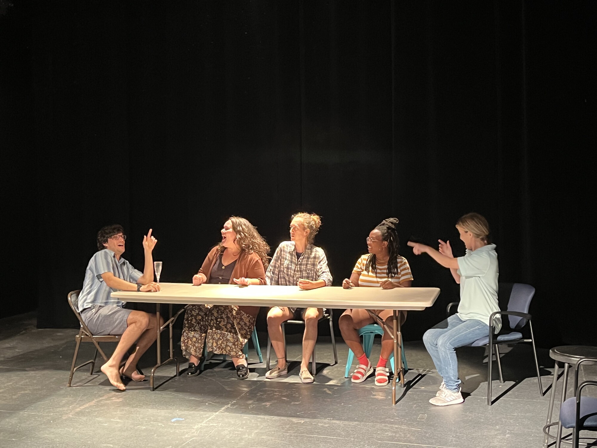 Five performers sit at a long folding card table on stage and laugh.