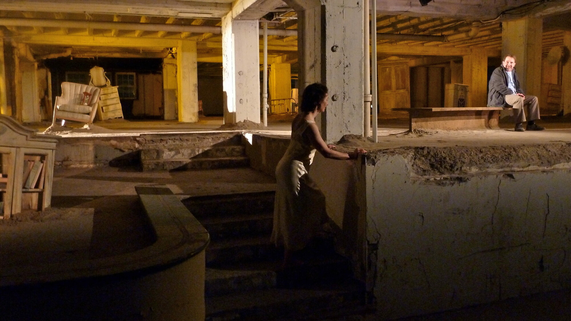 Two actors perform in a large cement room.