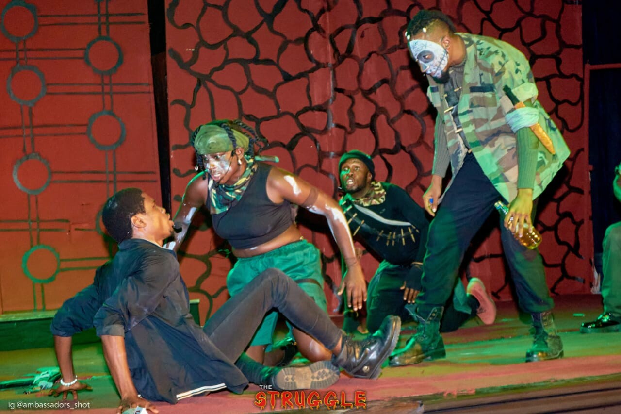 Two performers dressed as soldiers approach a third performer who is seated on the floor trying to move away from them.