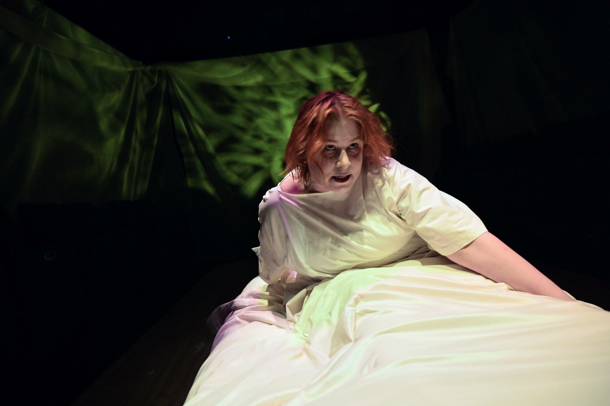 An actress sits up in bed under green lighting.