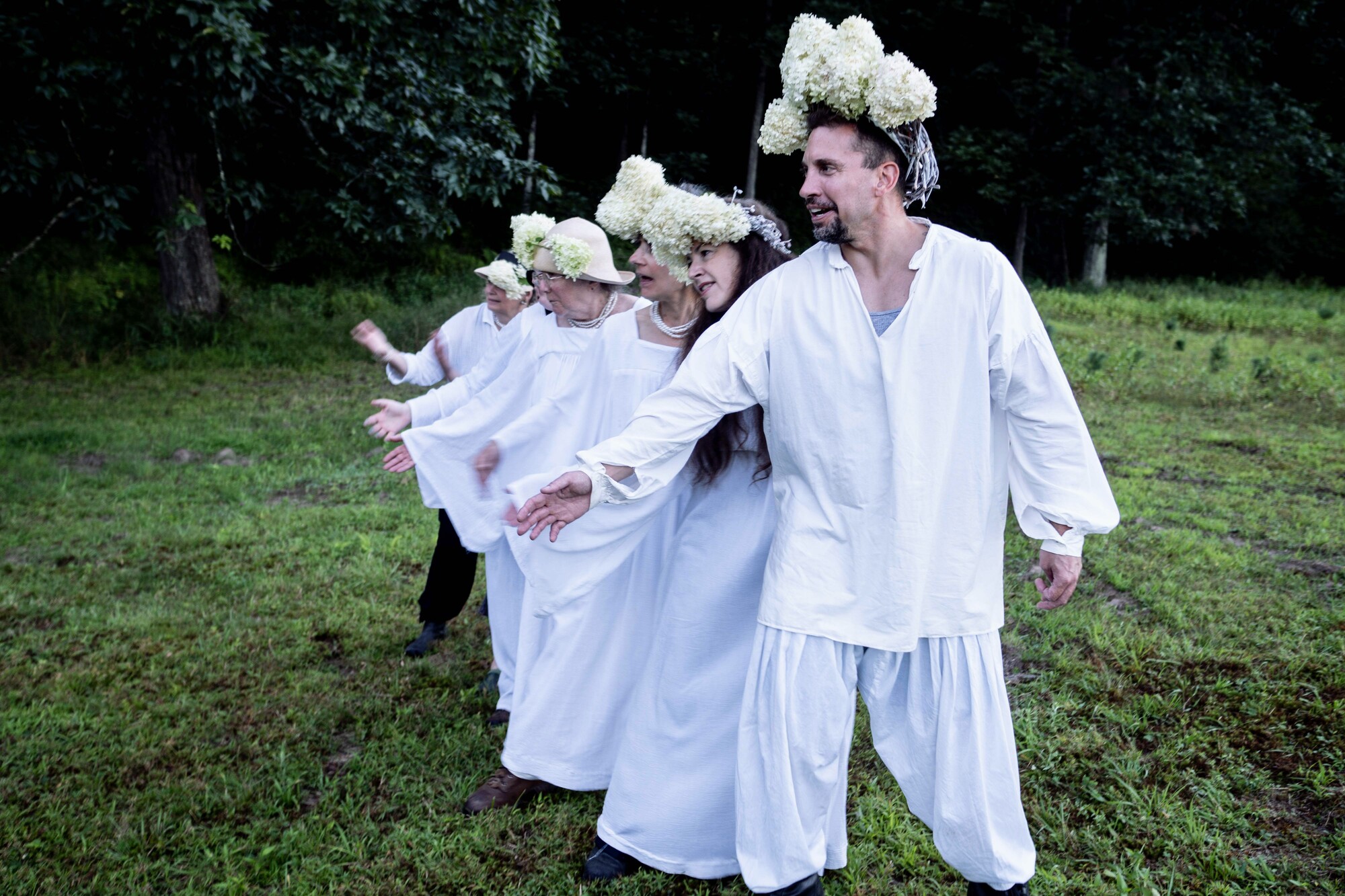 Performers in white with floral headpieces stand in a field.