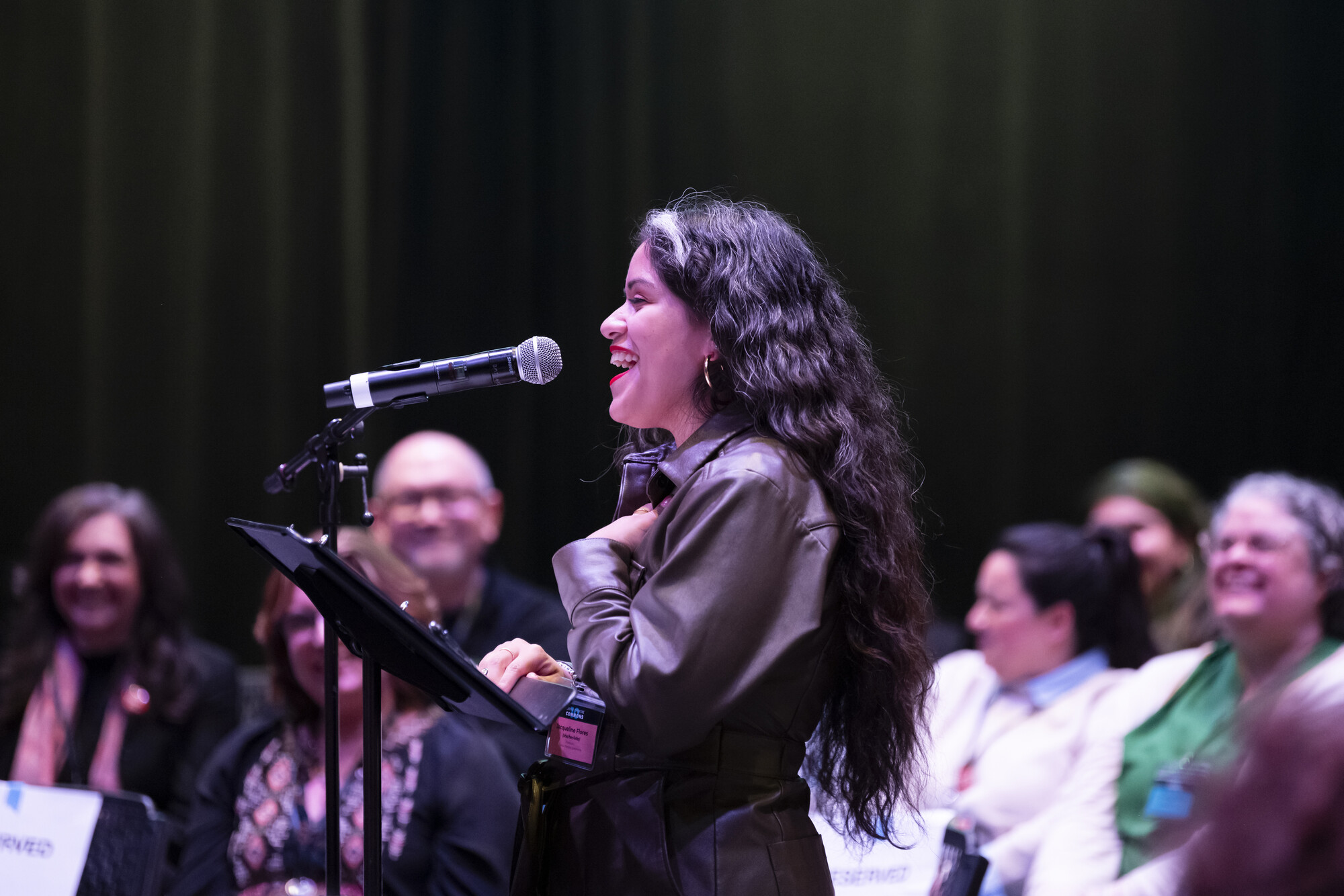 A woman with long dark hair stands in front of a microphone.