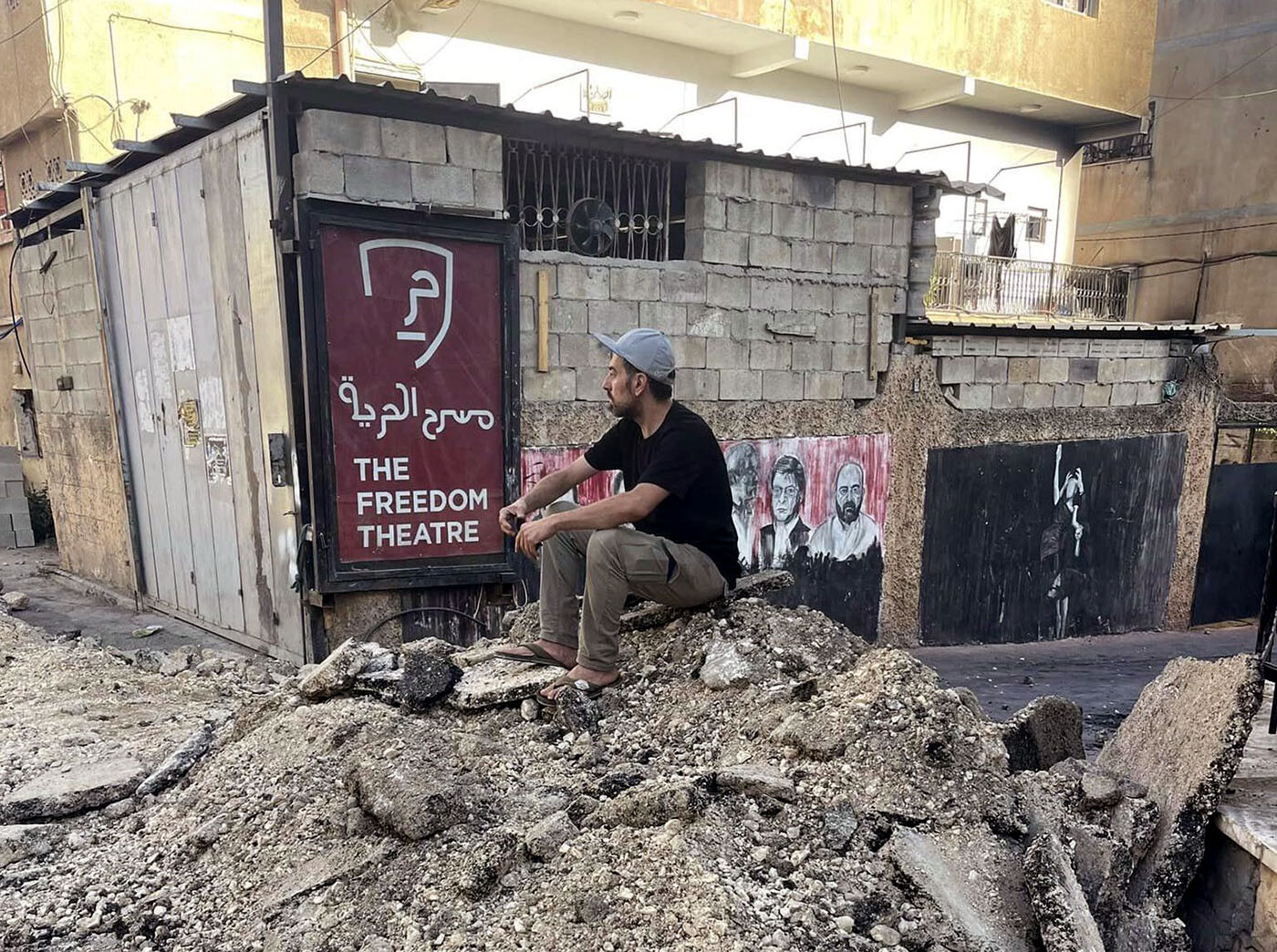 A man sits on a pile of rubble in front of a sign reading The Freedom Theatre.