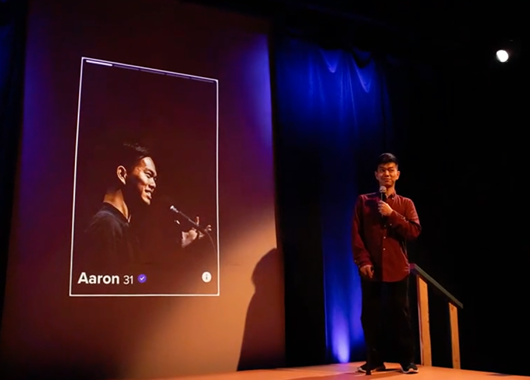 A man stands on stage in front of a projector screen of a dating app profile. 