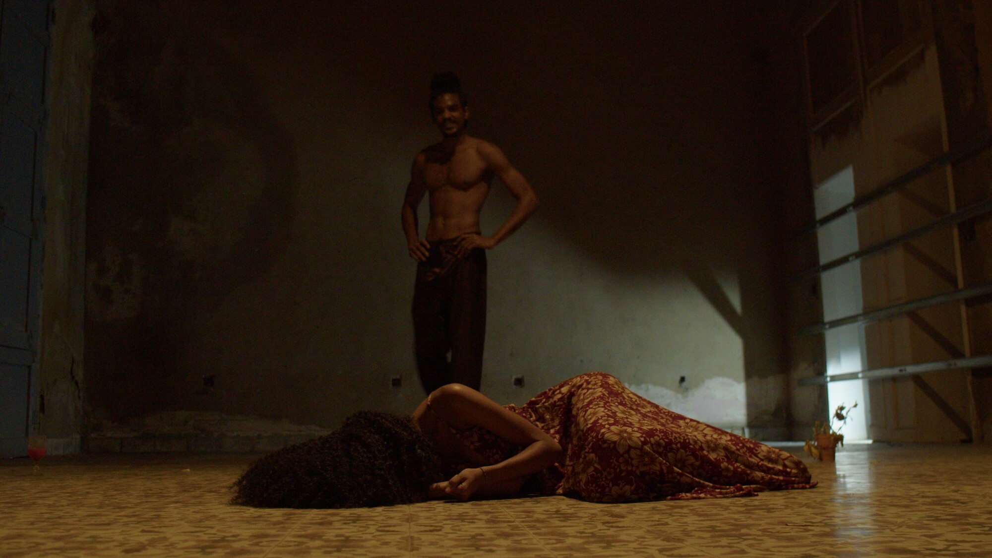 Two actors rehearse in a dimly lit space, with one standing and one laying on the ground.