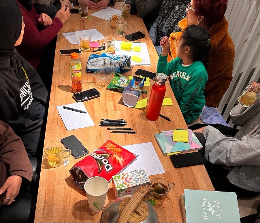 Woman sit around a workshop table covered in notebooks, paper and snacks.