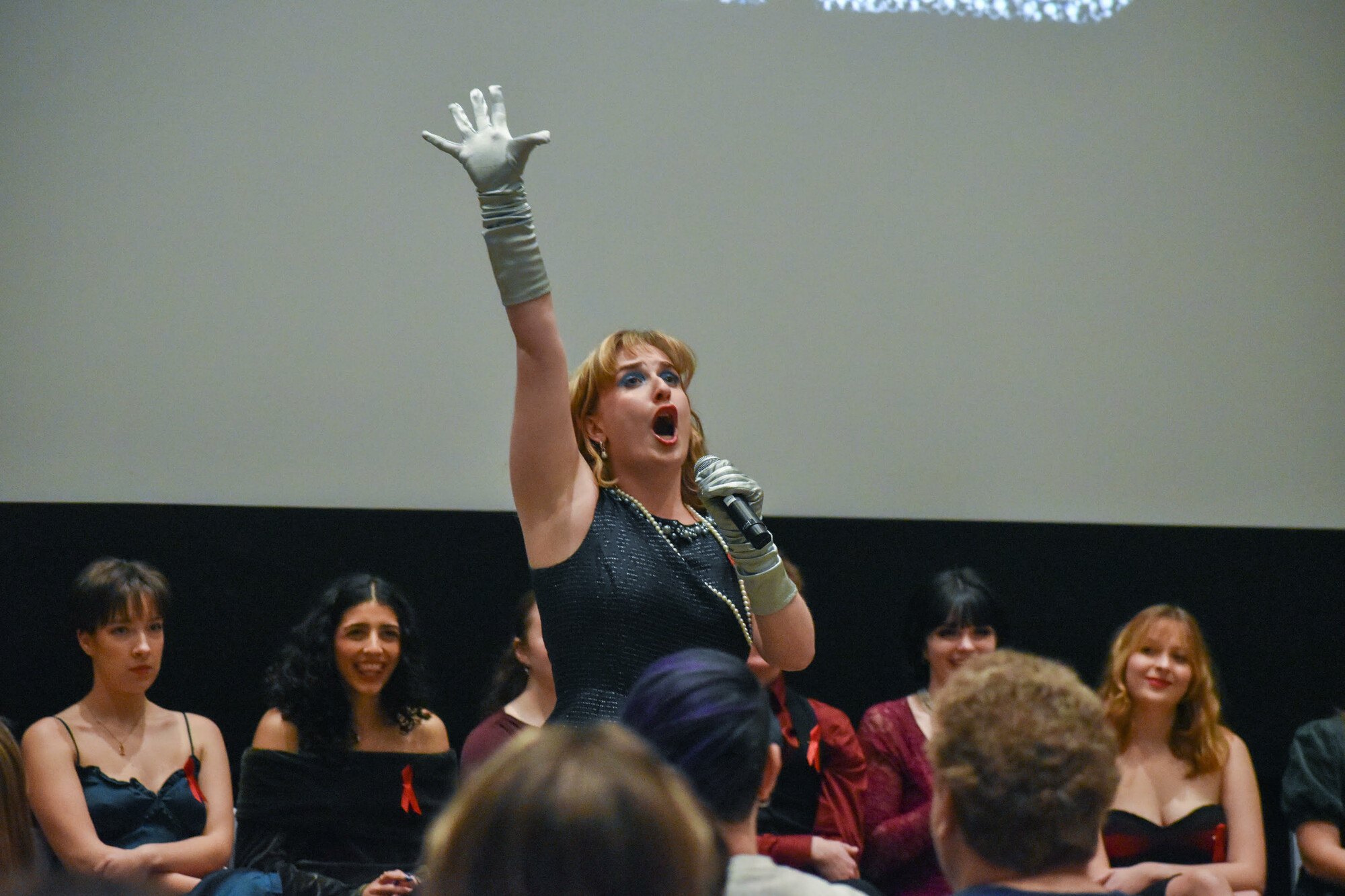 A soloist performs sings into a microphone in front of the rest of the cast of a caberet.