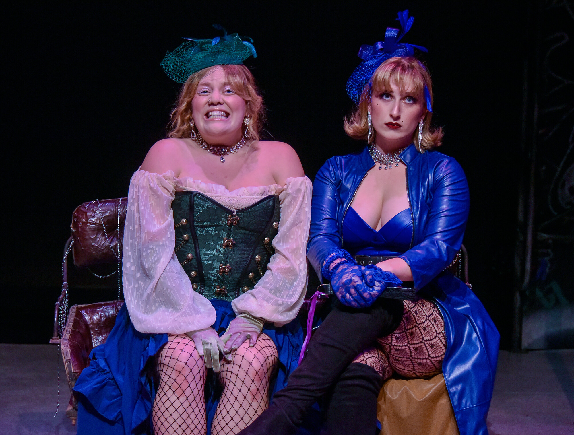 Two actors wearing brightly colored dresses sit on a leather bench onstage.