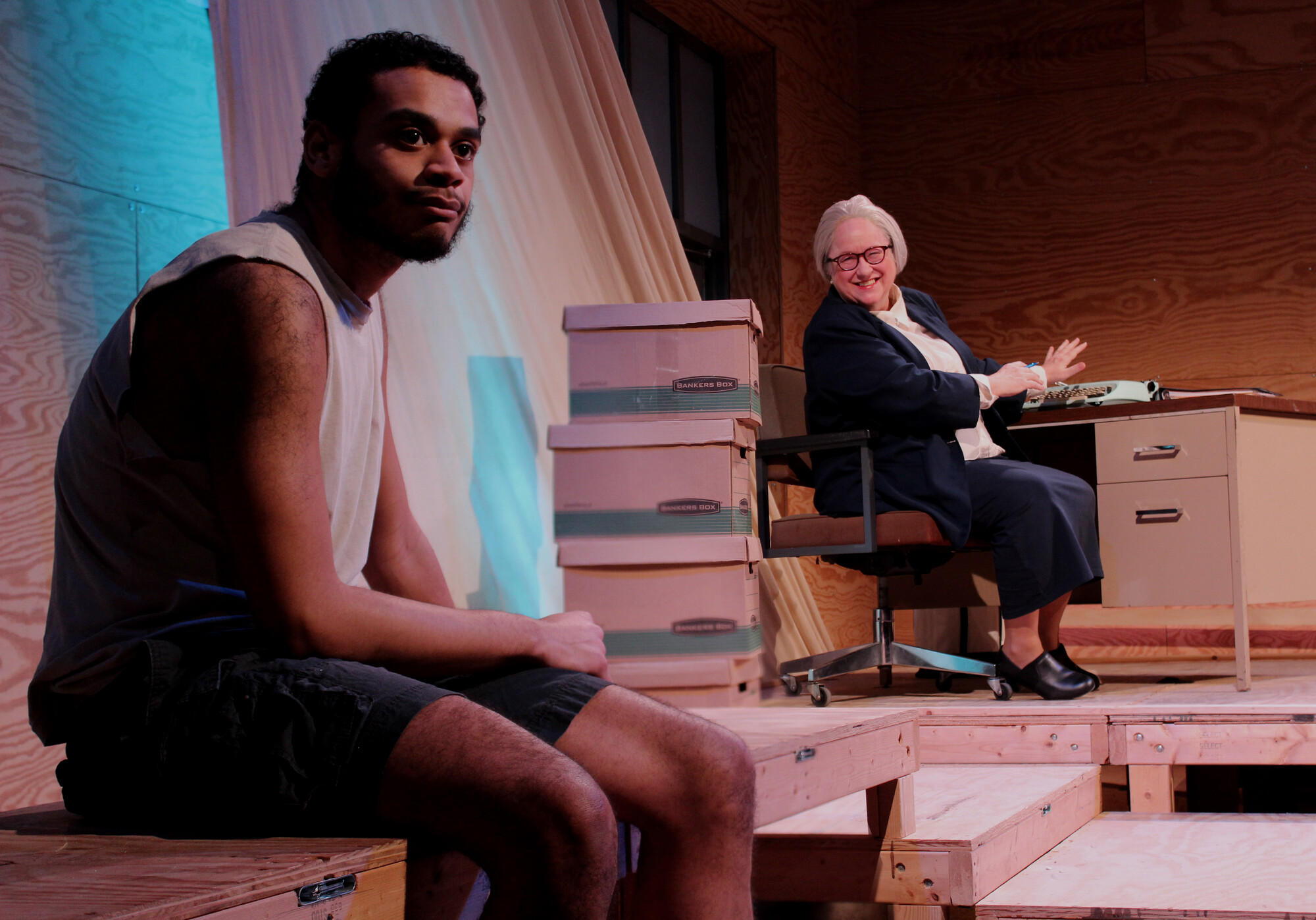 Two actors sit on wooden platforms surrounded by cardboard boxes on stage.