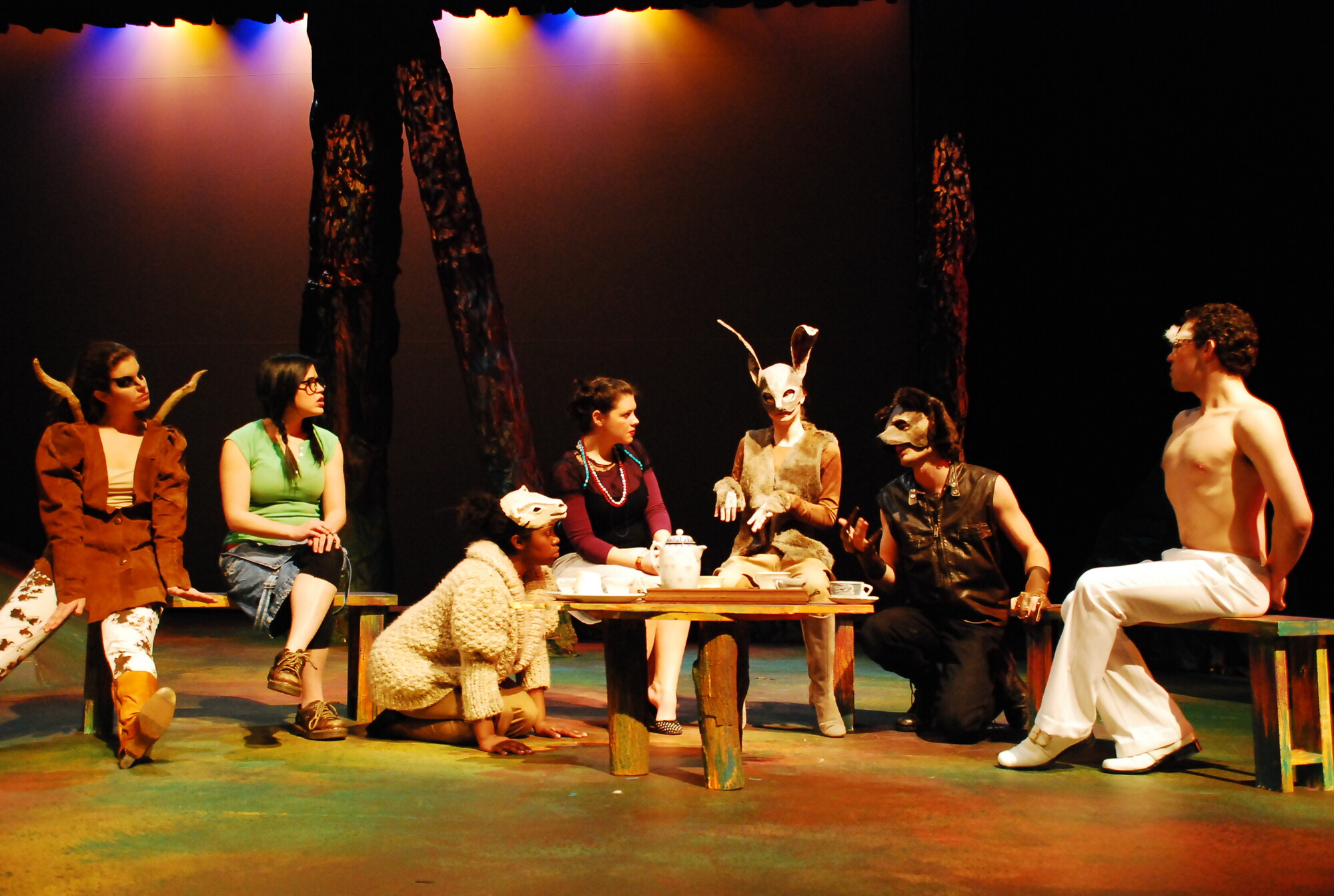 A group of actors in surreal costumes sit on benches on a colorful dimly lit stage.