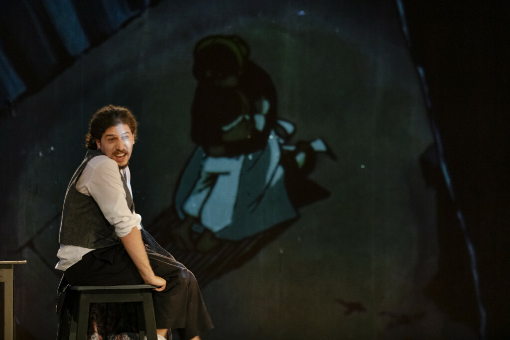 An actor sits on stage in front of a projected illustration.
