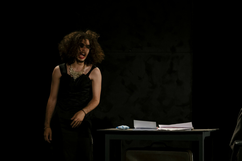 An actor stands on a dimly lit stage in front of a table.