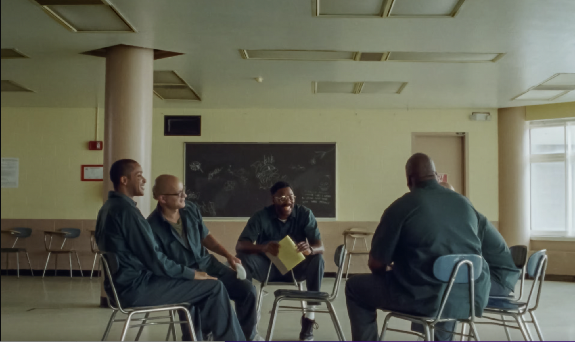 A still from the movie sing sing featuring actors sitting in a circle on metal chairs.