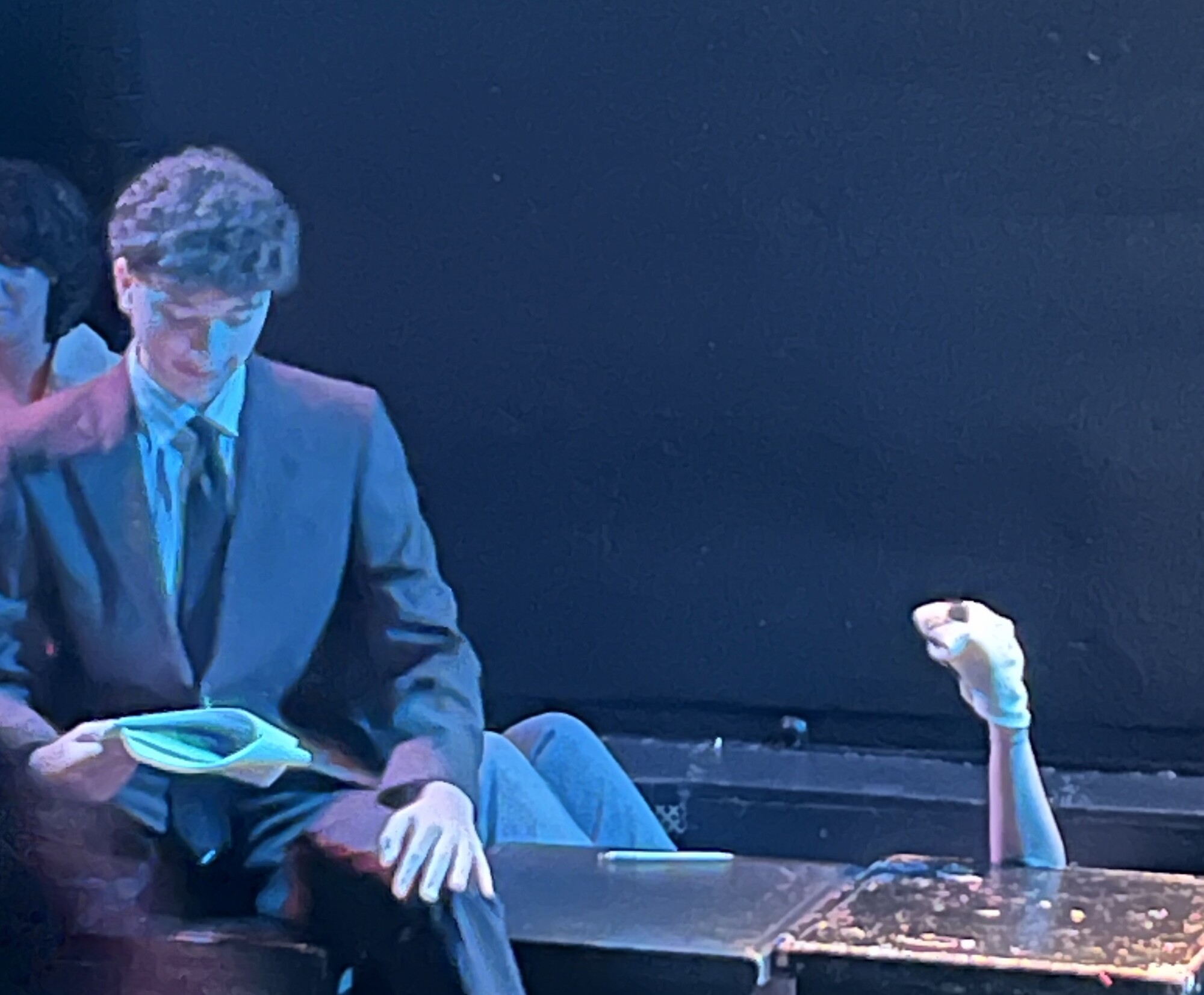 An actor sits on a cube next to another actor operating a sock puppet.