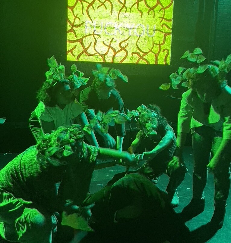 A group of actors in plant hats act in front of a projector onstage.