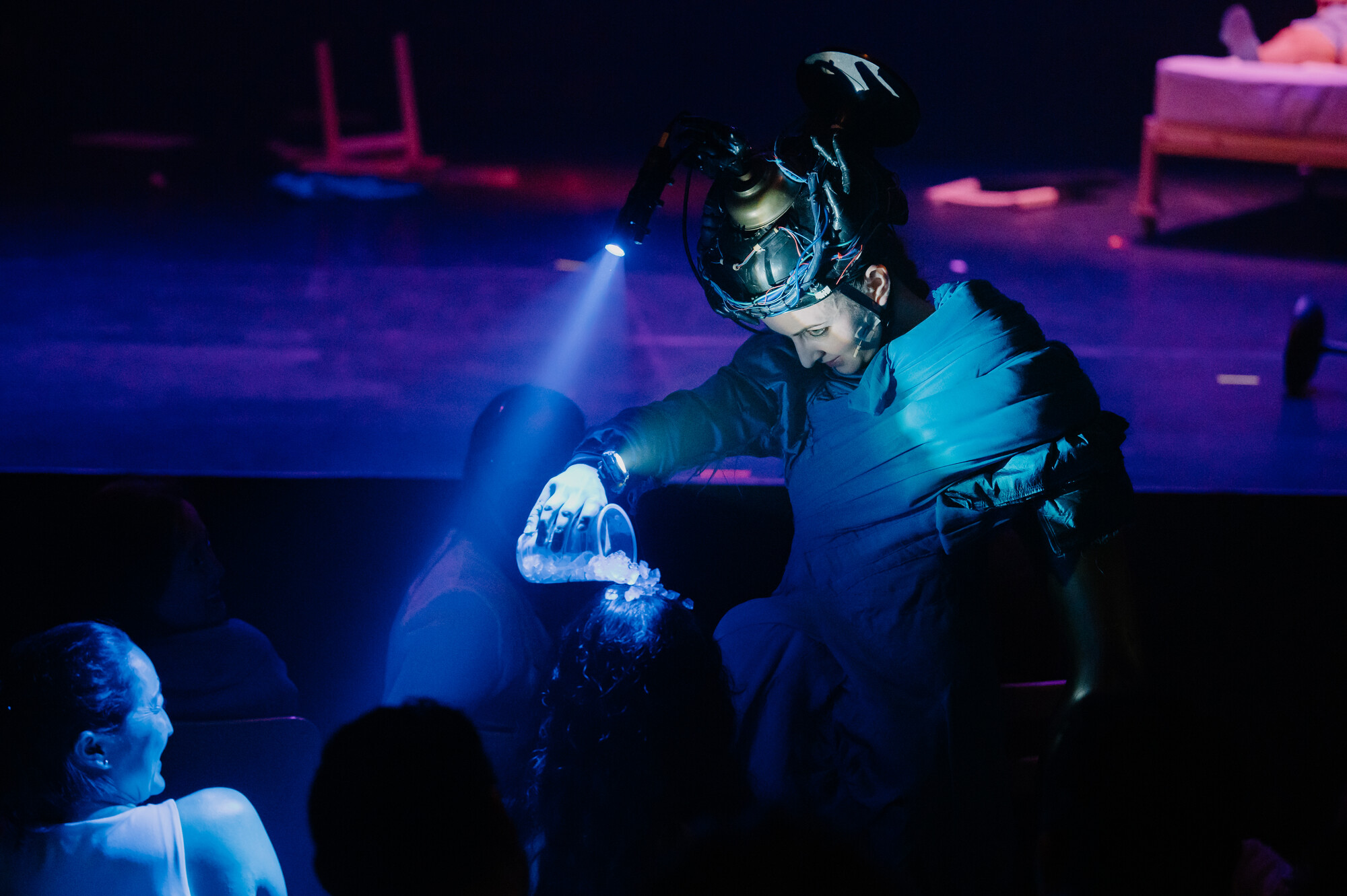A performer wearing a helmet with a flash lit does crowdwork on a dimly lit blue stage.