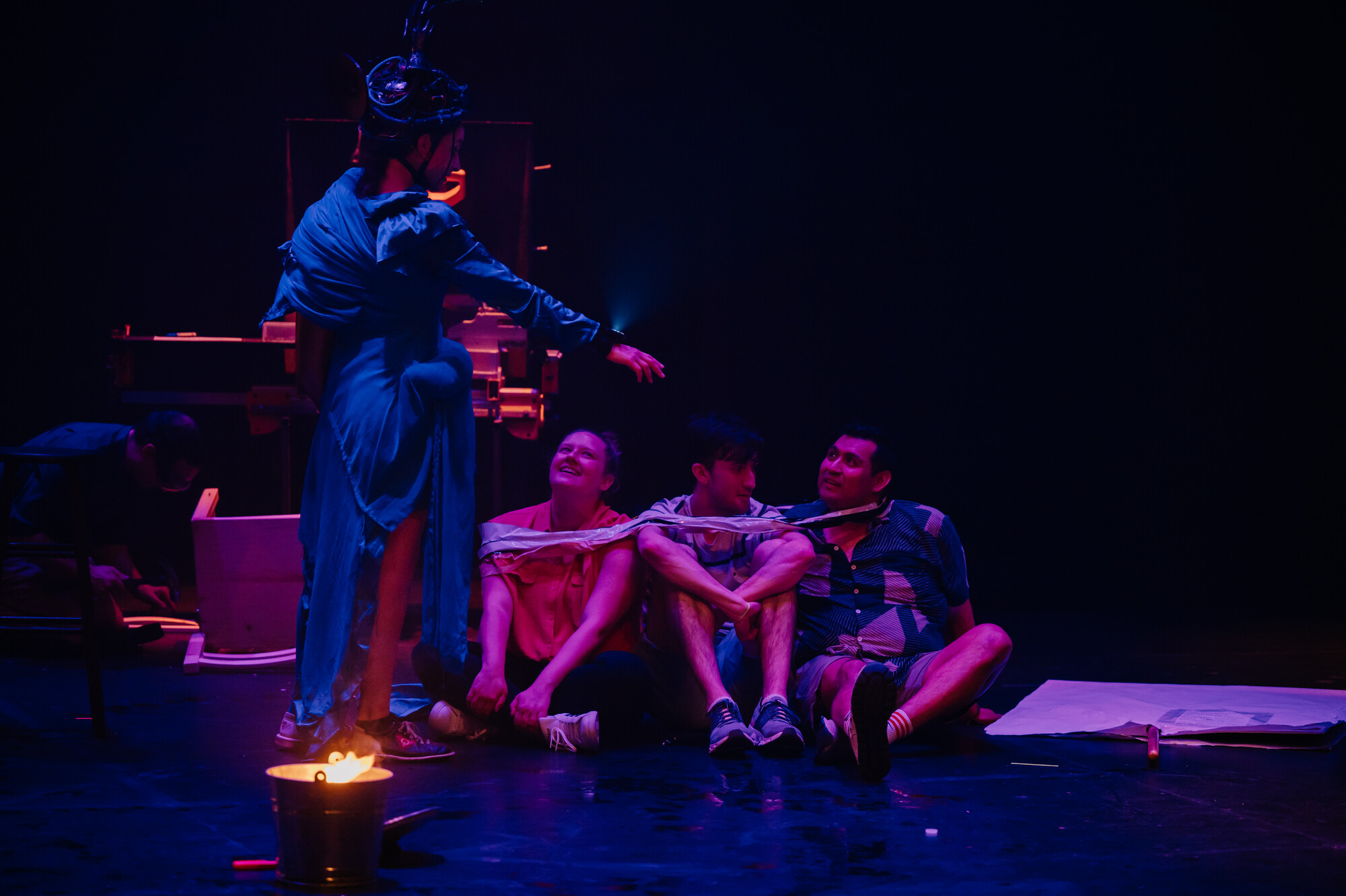 A performer stands, talking to three audience members sitting on the edge of a dimly lit stage. 
