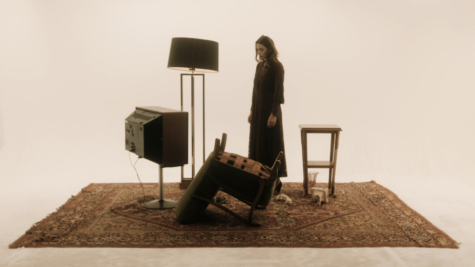 A woman standing on a rug surrounding by a lamp, tv, side table, and overturned chair. 