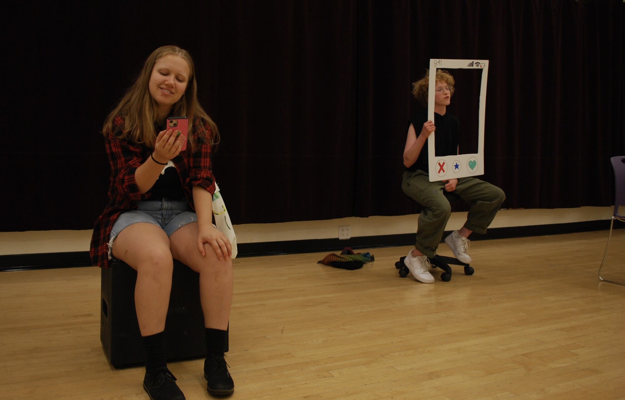 One person sits looking at a mobile phone in their hand and the other sits behind a cut out of a dating app frame. 