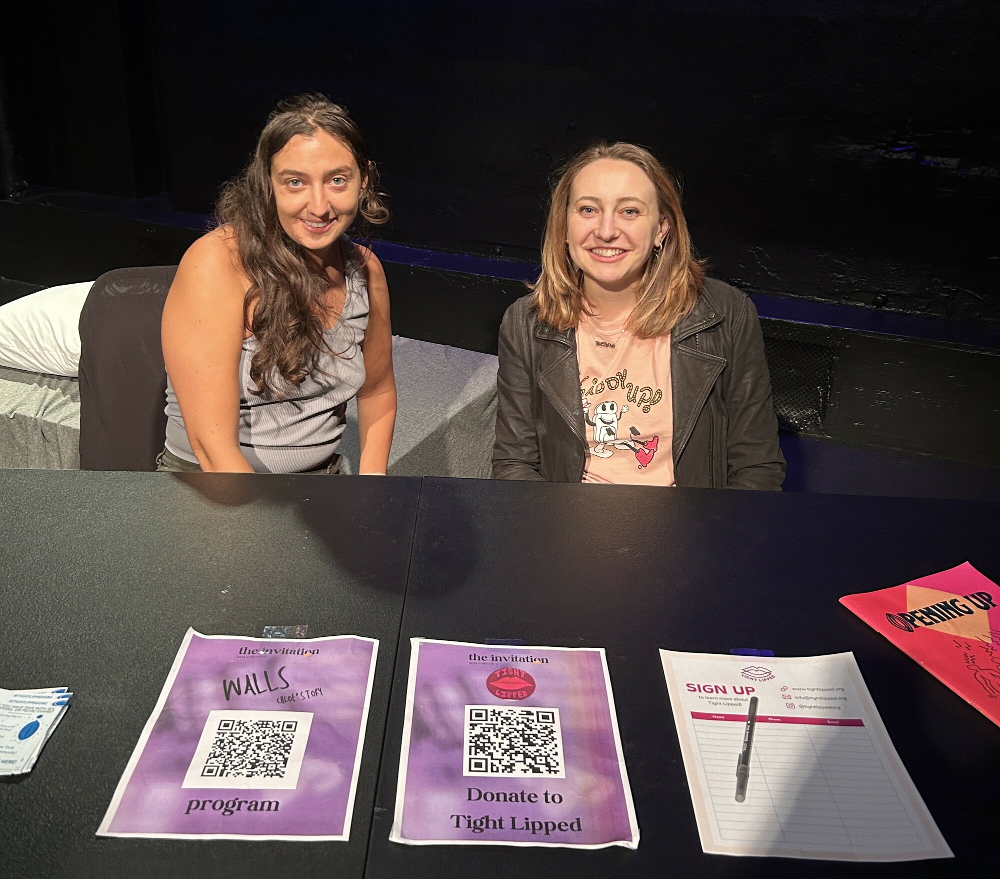 Two people sit behind a table with flyers laid out in front of them. 