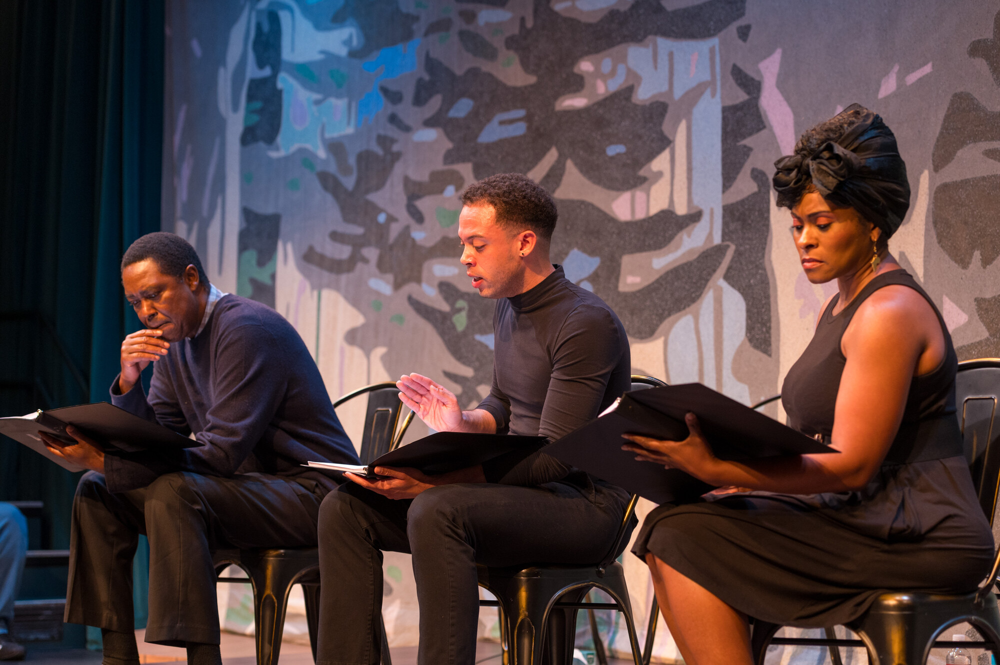 Three people in black clothes sit on stage reading from books in their hands. 