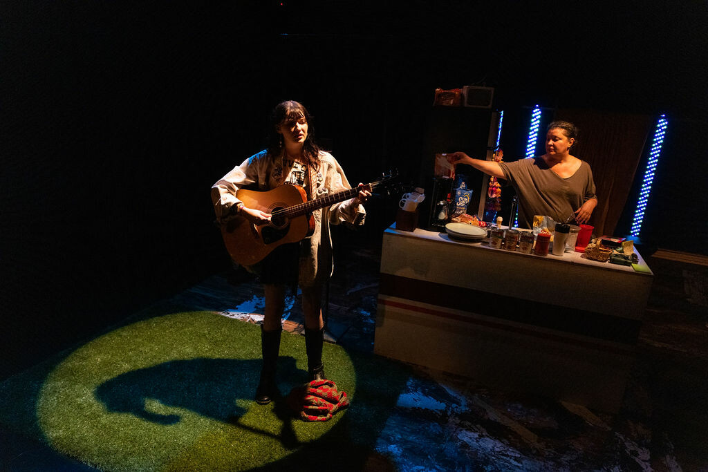 A person plays guitar on stage while another person is cooking in an on stage kitchen. 