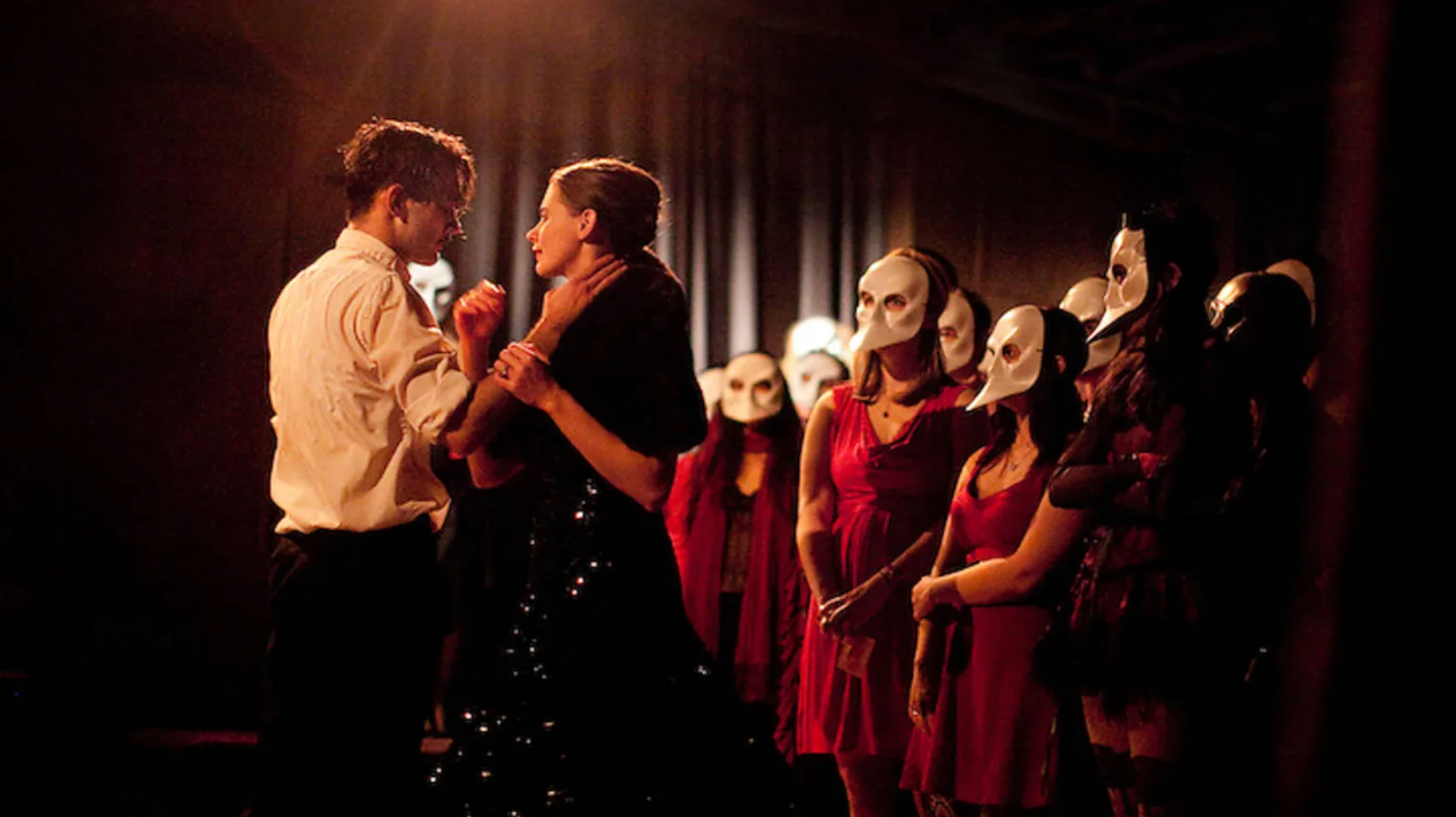 A man and a woman are in a passionate embrace and are watched by a group of people in white masks. 