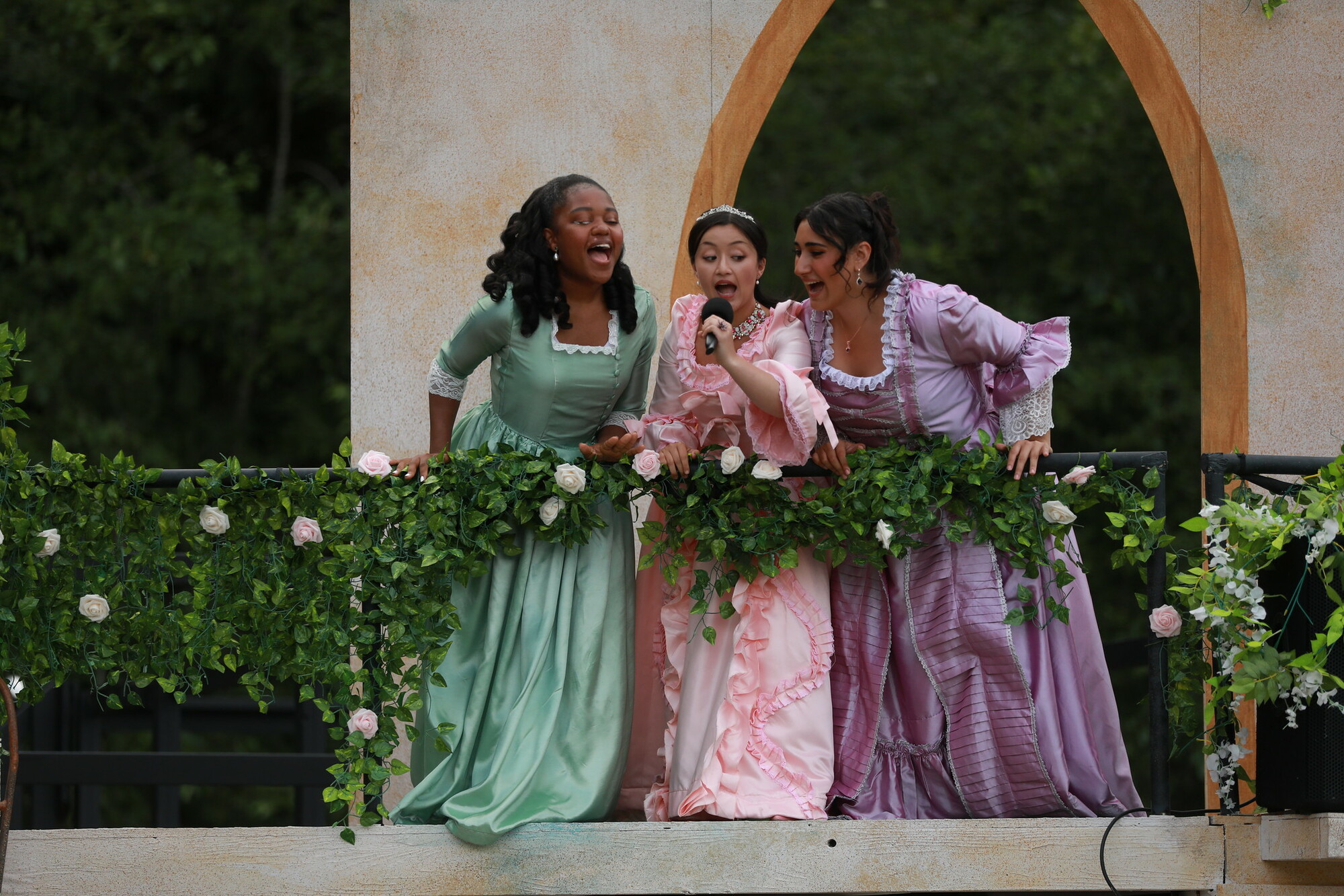 Three women in regency era dresses sing on a balcony.