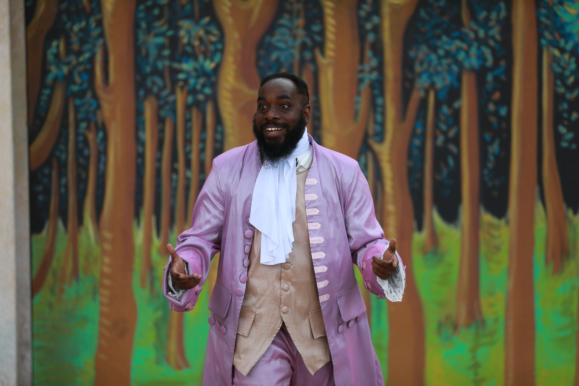 A man in a historical purple coat stands in front a forest backdrop.  