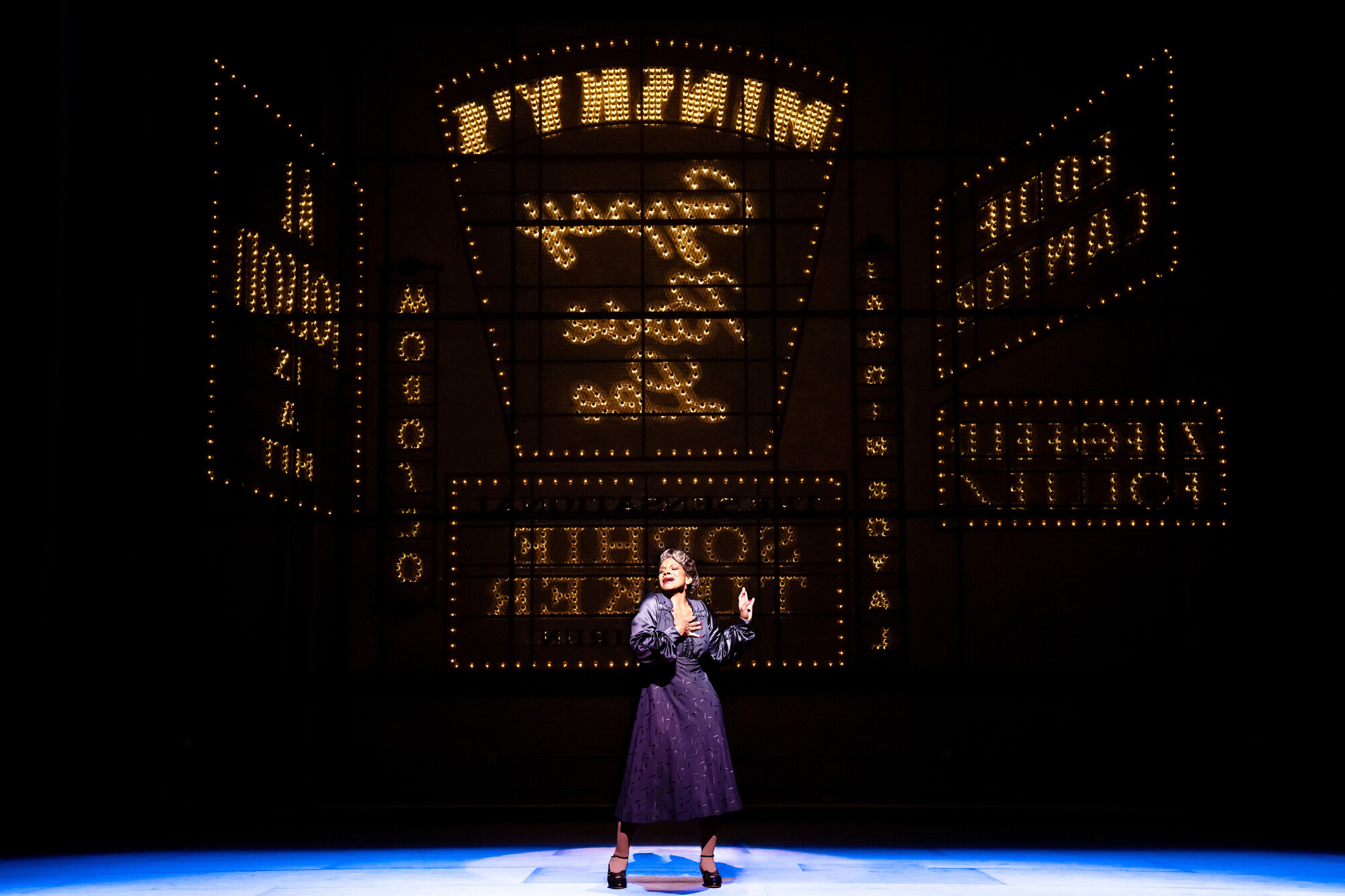 A woman in a purple dress stands center stage with bright signs behind her. 