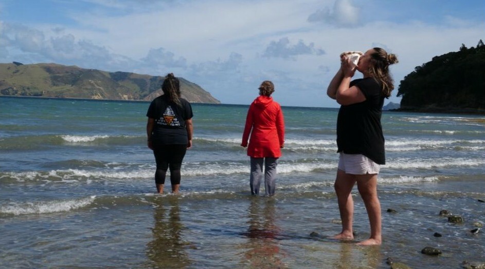 Two people stand in shallow water while a third person stands behind them blowing a conch. 