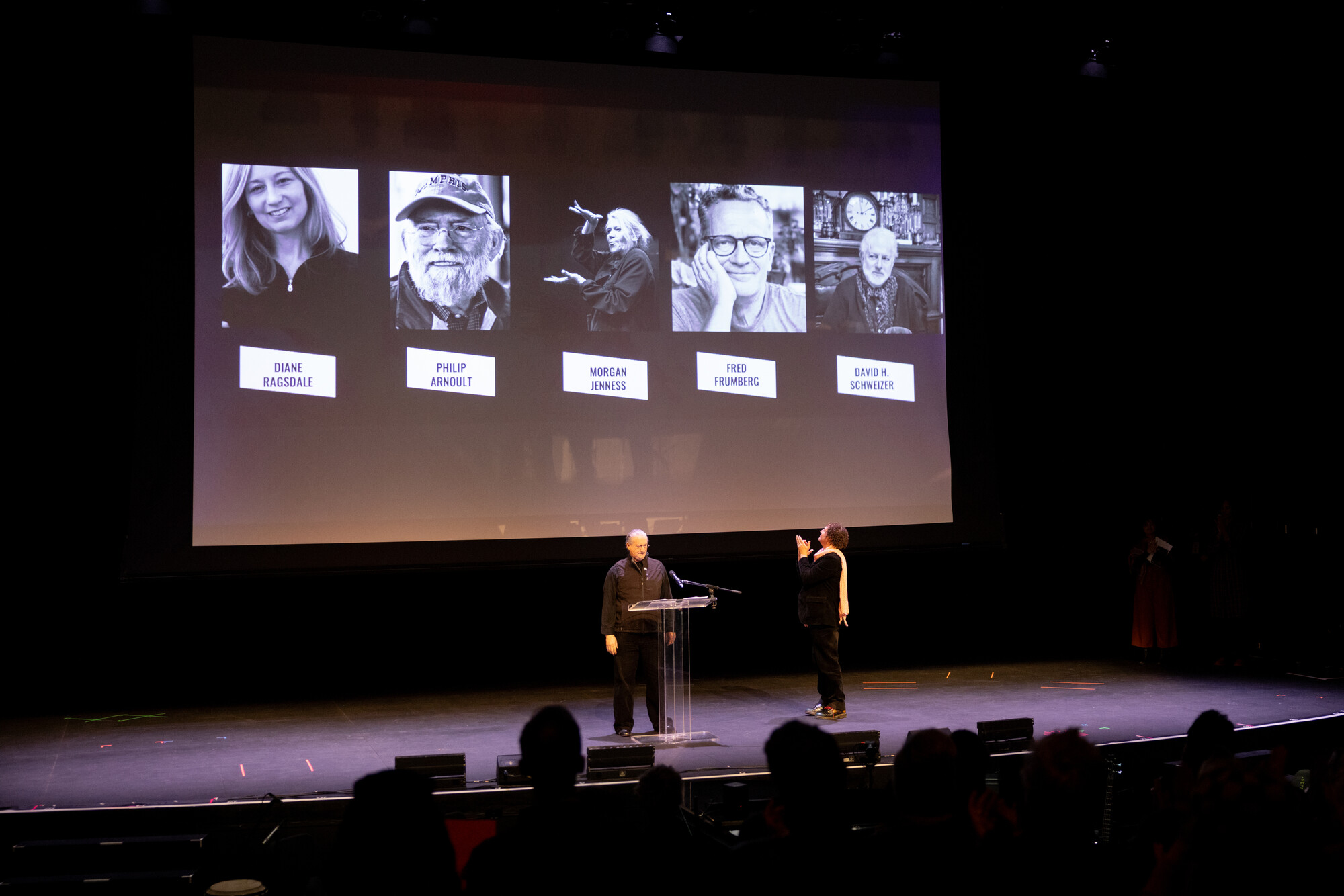 Two people on a stage with a projection behind them of headshots. 