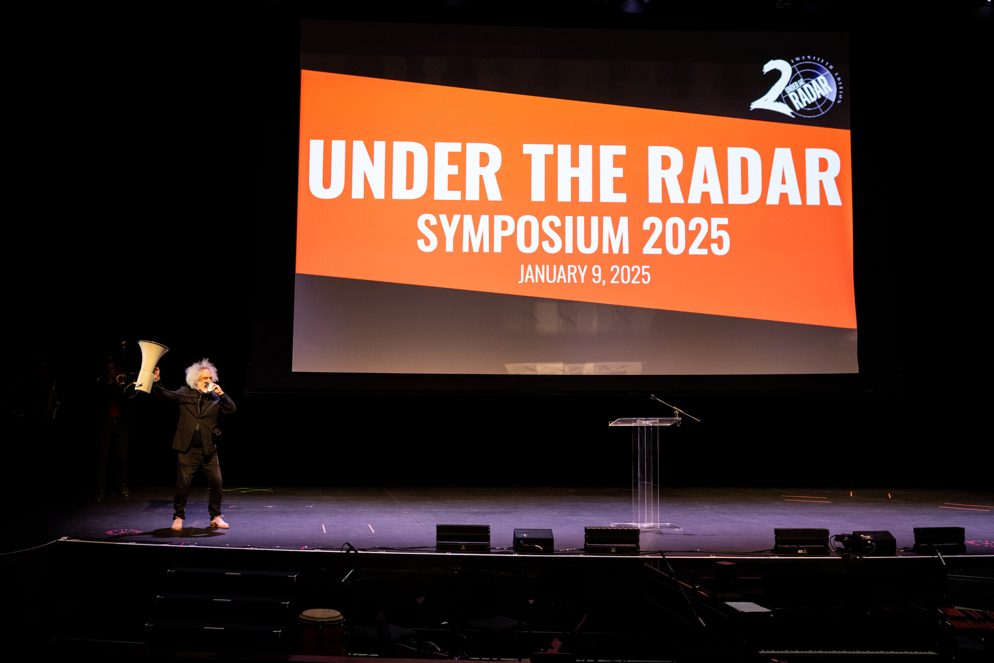 A person on stage with a mega phone and a screen that reads Under the Radar Symposium 2025.