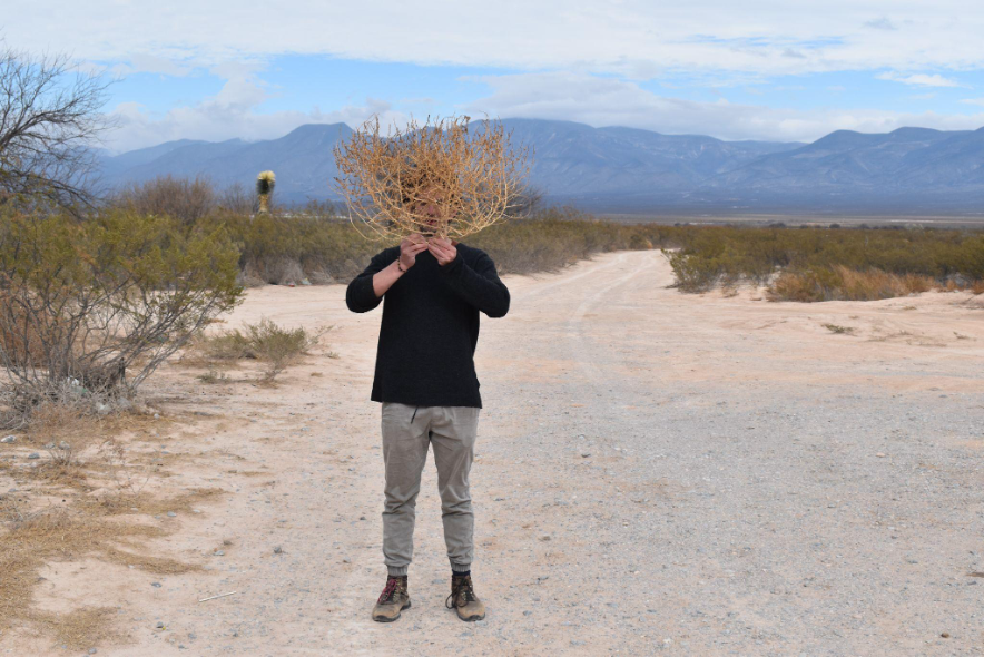 A person standing in a desert holding a tumbleweed in front of their face.