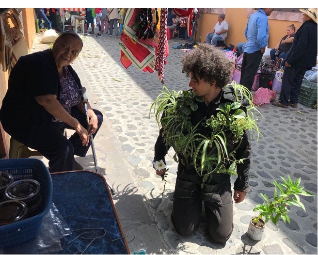 A man dressed in all black with plants on his clothes kneels on a street.