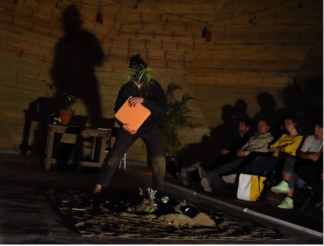 A person performing in dark lighting with plants on their head and holding orange papers.