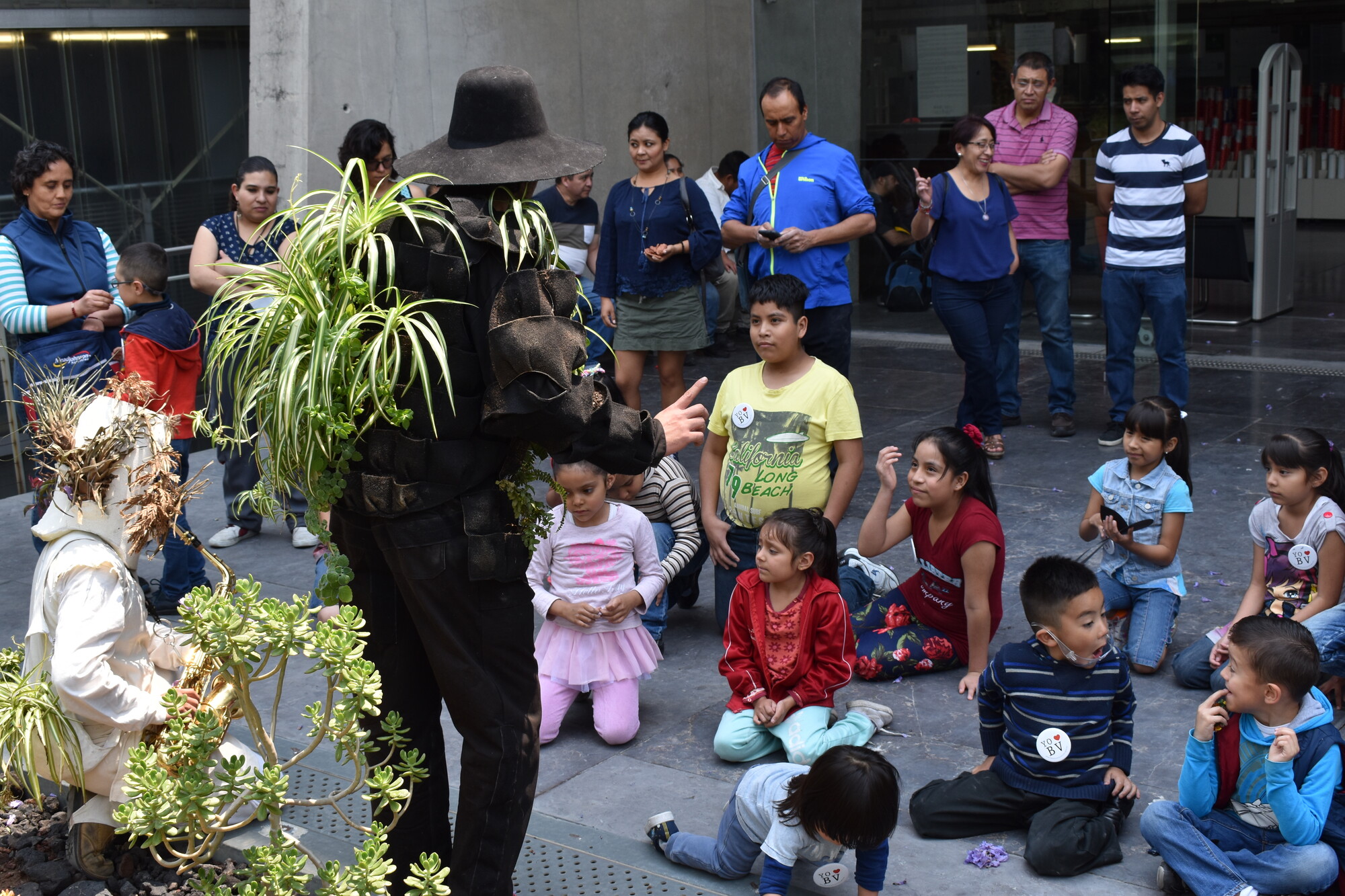 A man in all black and covered with plants addresses a crowd of children.