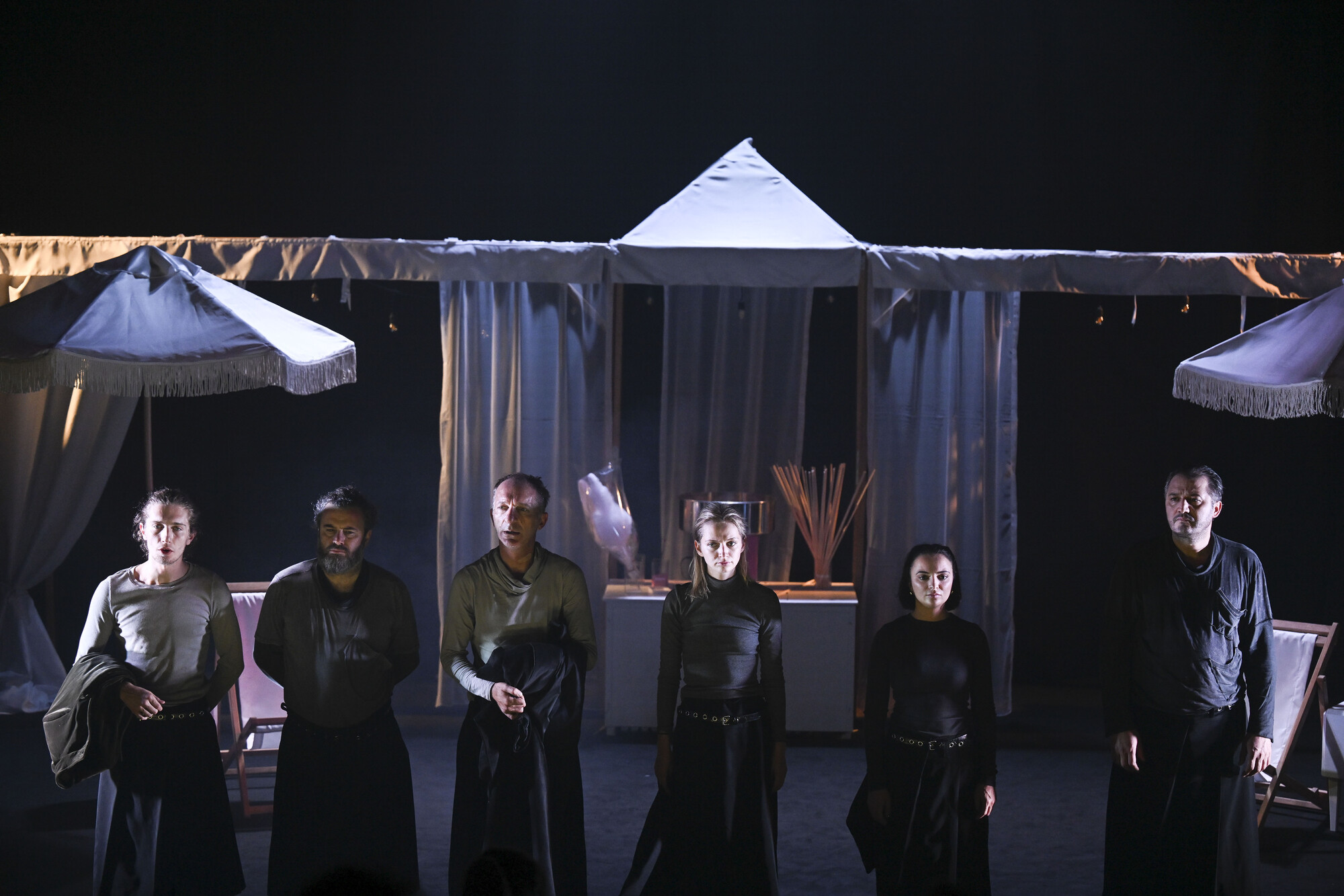 A group of people in dark clothing stand in a line on stage with grey umbrellas behind them.