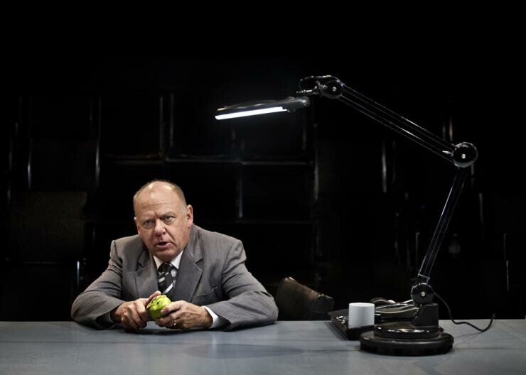 A man in a suit sitting at a desk and peeling an apple.