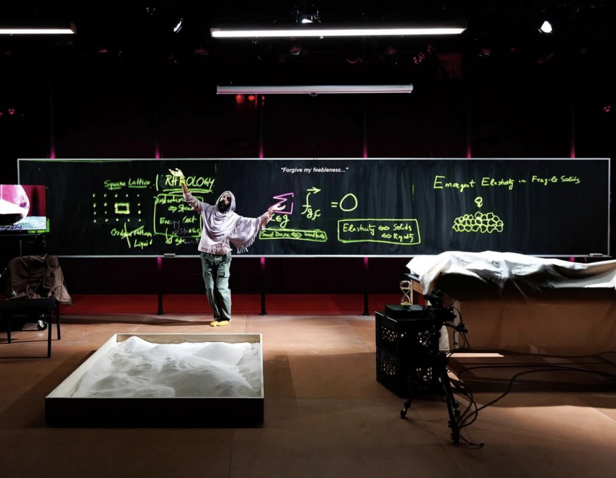 A person in a shawl standing in front of a black board with neon green writing on it.