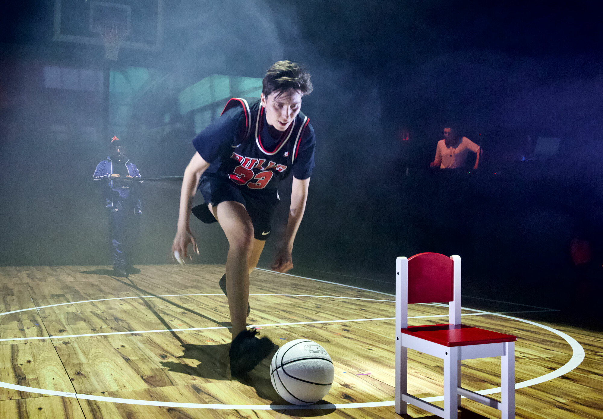 A person in a basketball uniform bending down for a ball near a small chair.