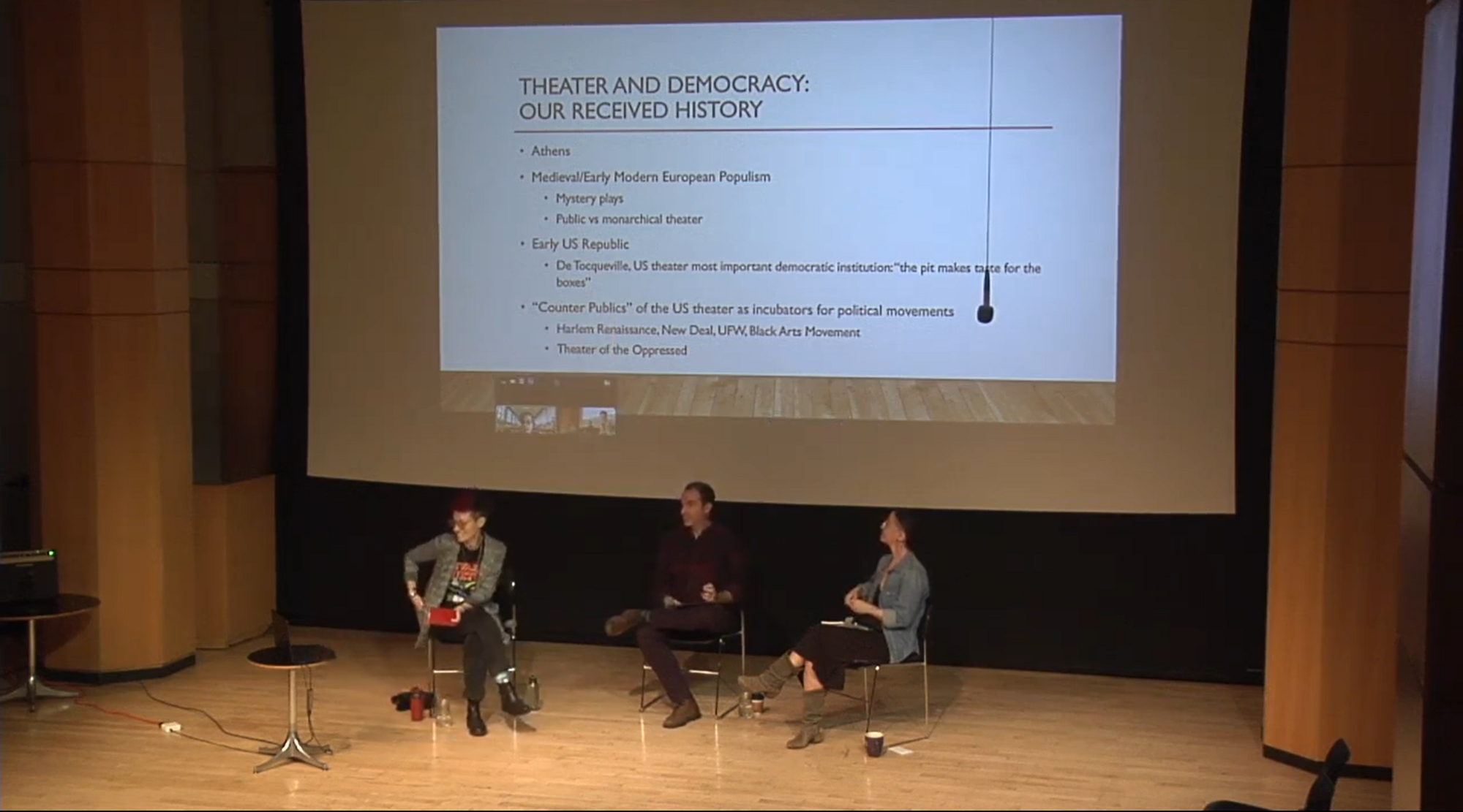 A panel of three people sitting on a stage with informational text on a screen behind them.