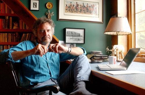 A man in a blue button up shirt and jeans sitting at a desk.