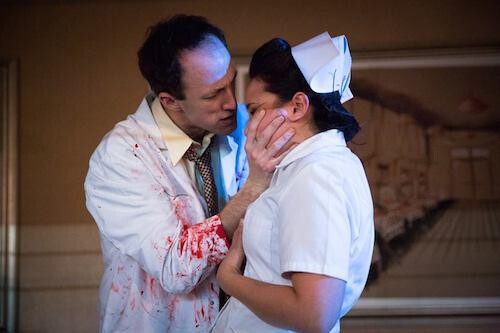 A man on stage dressed in a bloody doctor's coat grabbing the face of a woman dressed as a nurse.