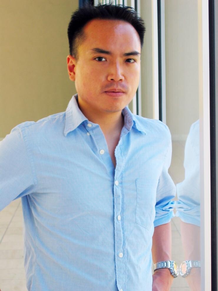 An Asian man in a blue button up looking seriously at the camera. 